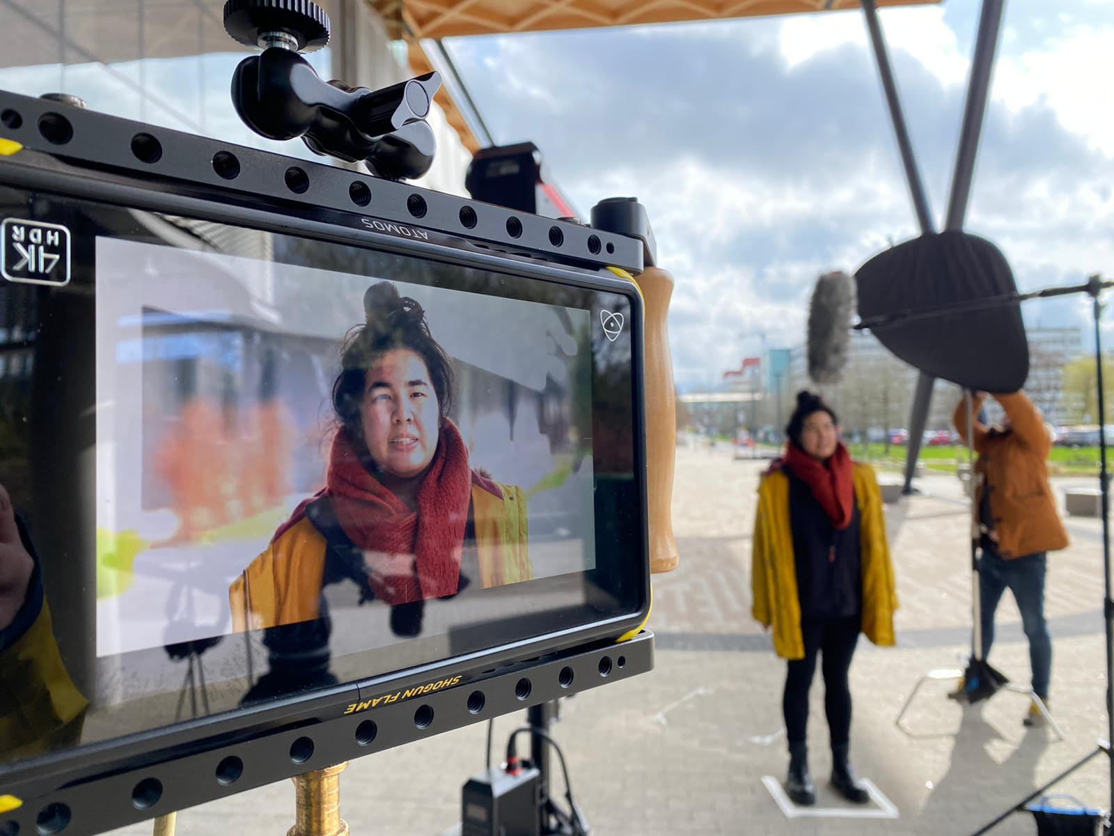 A woman stands being interviewed with the camera mirroring this in the far left of the picture.